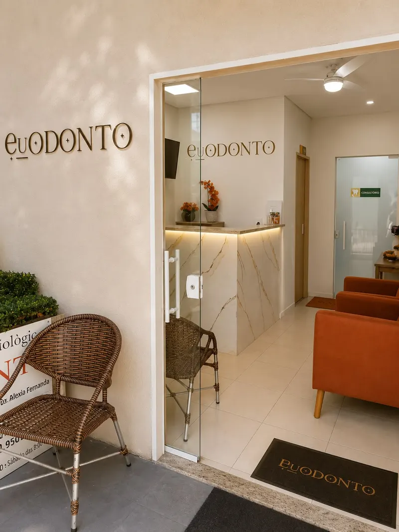Recepção da EuOdonto: entrada de vidro com balcão de mármore, letreiro da clínica na parede e sofás terracota